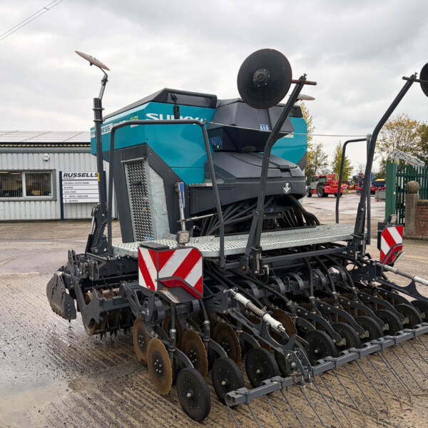 Large blue-green disc cultivator on a trailer in a yard beside a grey metal building.