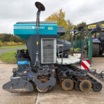 Large blue farming machine with disc blades and rollers parked on a paved surface, a blue tractor in the background near a green building.