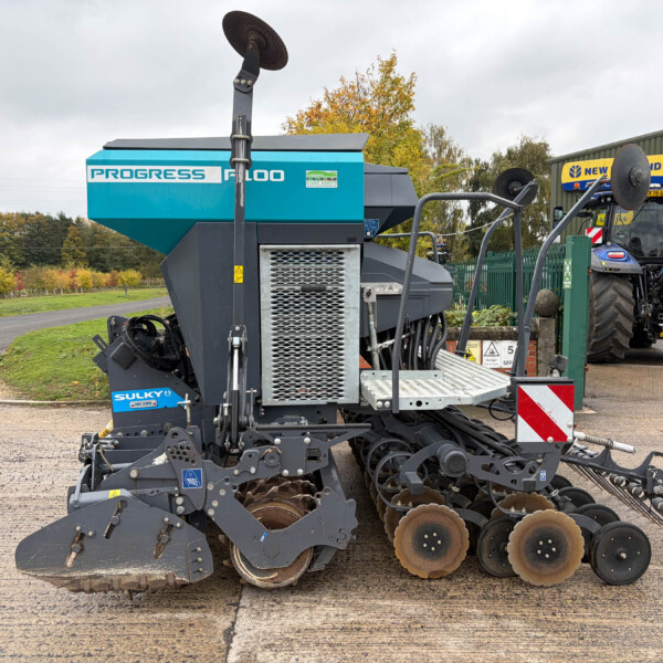 Large blue farming machine with disc blades and rollers parked on a paved surface, a blue tractor in the background near a green building.