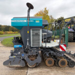 Blue agricultural seed drill with large round discs attached to a metal frame, parked by a New Holland tractor in a driveway showroom area.