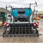 Front view of a large, multi-row tine harrow attached to a farming trailer, with a blue Sulky machine in the background in an industrial yard.