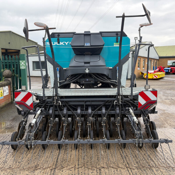Front view of a large, multi-row tine harrow attached to a farming trailer, with a blue Sulky machine in the background in an industrial yard.