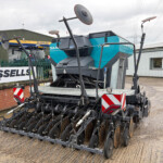 Large blue and gray agricultural seeding machine in a yard, with 'Russells' and 'New Holland Agriculture' signs in the background.