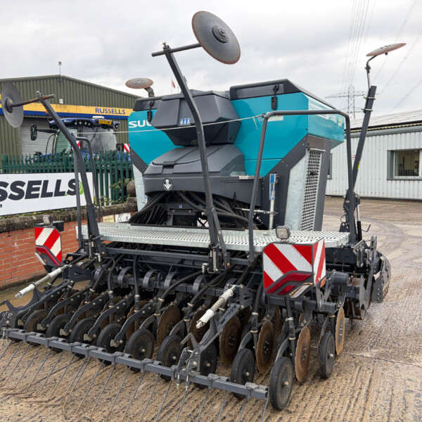 Large blue and gray agricultural seeding machine in a yard, with 'Russells' and 'New Holland Agriculture' signs in the background.