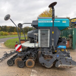 Large agricultural planter with multiple disk rows for sowing, parked on a paved surface near a blue Progress machine and a New Holland Agriculture sign in the background.