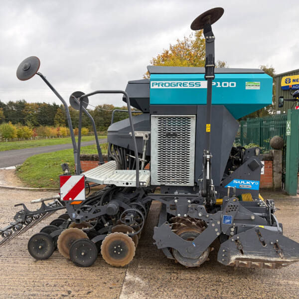 Large agricultural planter with multiple disk rows for sowing, parked on a paved surface near a blue Progress machine and a New Holland Agriculture sign in the background.