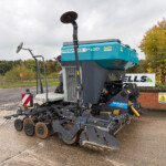 Progress P100 teal and gray agricultural harvester parked outdoors in a yard, with autumn trees in the background.