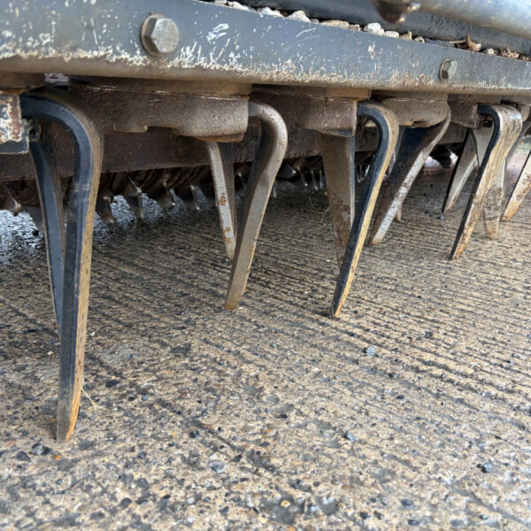 Close-up of rusted metal tines on a cultivator beneath a weathered frame, with a muddy gravel ground beneath.