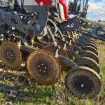 Close-up of a rusted disc harrow attached to a farming implement, showing multiple circular metal discs and gray framework on rocky soil.