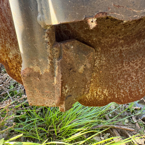 Close-up of a rusted metal joint with peeling paint and dry vegetation around its base.