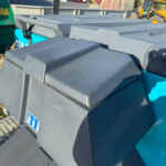 Row of dark gray outdoor waste bins with ridged lids, set near green industrial machinery in a construction yard, with a yellow vehicle in the background