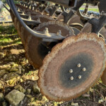 Close-up of rusted circular cutting discs and bolts on a farm planter, with rocky ground in the background