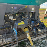 Close-up of a farm machine's mechanical components: hoses, gauges, and metal framework with a green hopper above; field in distance.