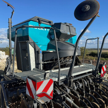 Blue agricultural seeder machine with disc implement and red-and-white safety panels on a gravel site under a clear blue sky.