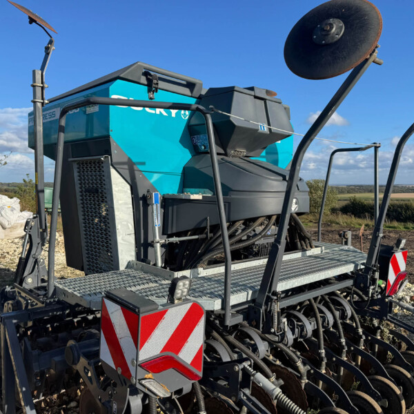 Blue agricultural seeder machine with disc implement and red-and-white safety panels on a gravel site under a clear blue sky.