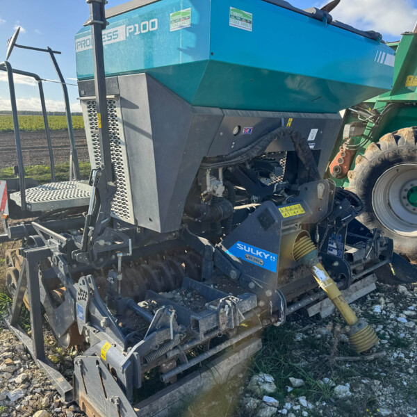 Blue hopper and metal framework of a tractor-mounted harvester in a farmland field, with a large rear wheel visible and rocky ground nearby, under a blue sky.