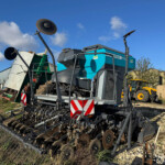 Large blue agricultural machine with circular discs and metal arms, parked on a gravel lot under a clear blue sky.