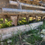 Close-up of muddy, rusted metal rollers and frame from a farm implement, with grass growing beneath.