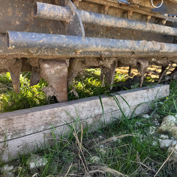 Close-up of muddy, rusted metal rollers and frame from a farm implement, with grass growing beneath.