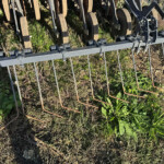 Metal agricultural implement with circular rusty rollers and a gray frame on soil amid weeds in a field setting.