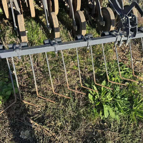 Metal agricultural implement with circular rusty rollers and a gray frame on soil amid weeds in a field setting.