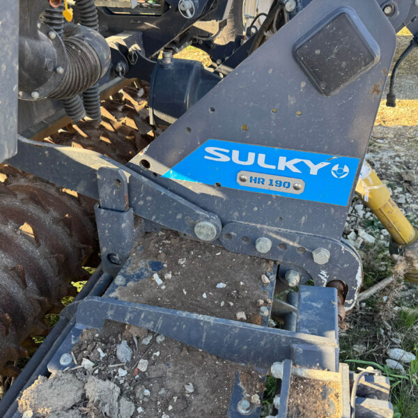 Close-up of a Sulky HR 190 farming implement showing a dirty metal frame, mechanical linkages, and a yellow hydraulic shaft on the right side.