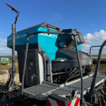Heavy construction machine with a blue-green body and metal steps, outdoors on a dirt site under a blue sky.