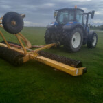 Blue tractor attached to a yellow cultivator with large spiked rollers on a grassy field under a cloudy sky.
