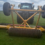 Blue tractor with an orange-yellow farming implement attached, wheels elevated off the ground in a grassy field under a cloudy sky.