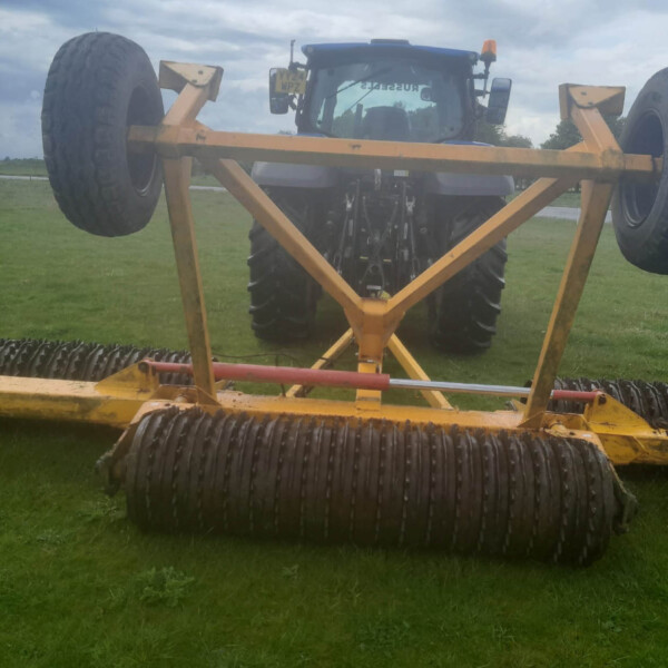 Blue tractor with an orange-yellow farming implement attached, wheels elevated off the ground in a grassy field under a cloudy sky.
