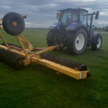 Blue tractor attached to a yellow cultivator with large spiked rollers on a grassy field under a cloudy sky.