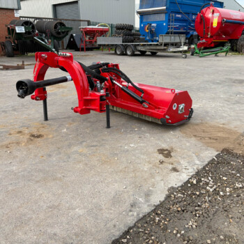 Red farming implement with hydraulic arms in the foreground on a concrete yard, with industrial machinery and buildings in the background.