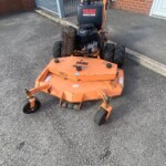 Orange SCAG commercial mower attached to a small utility tractor in a paved lot by a brick building.