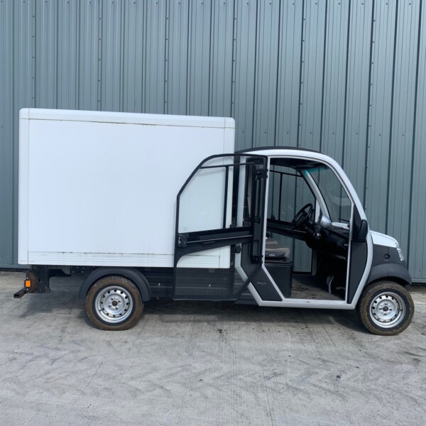 White delivery van with the driver’s side door open, parked against a tall gray corrugated metal wall.