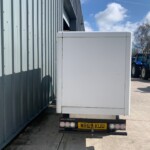 Rear of a white box trailer parked beside a blue corrugated metal building, with farm tractors visible in the distance on a sunny day.