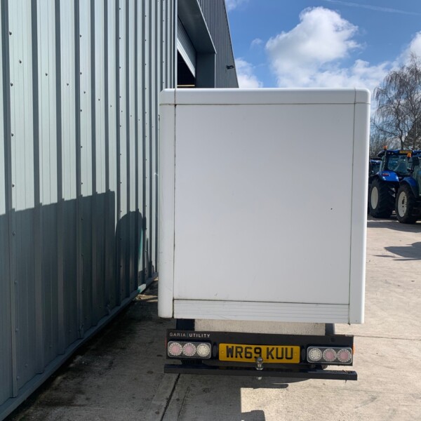 Rear of a white box trailer parked beside a blue corrugated metal building, with farm tractors visible in the distance on a sunny day.