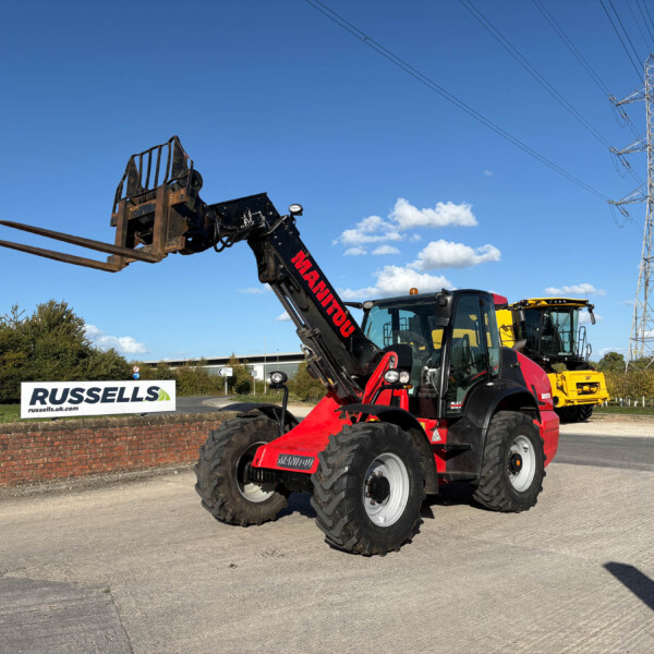 Red construction forklift with extended forks raised outdoors near a Russell sign and a power-line pylon against a blue sky.