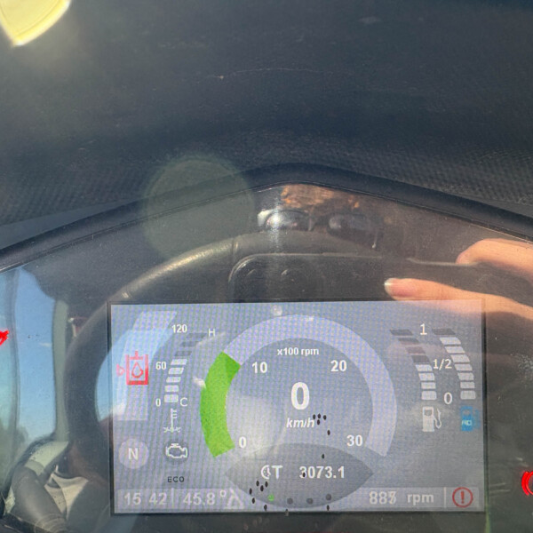 Close-up of a car's digital instrument cluster displaying speed (km/h), RPM, and warning icons including parking brake; dashboard reflected in the window.