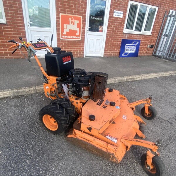 Orange SCAG lawn mower with exposed engine parked on the pavement in front of a brick building with reception signs.