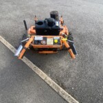 Small orange plate compactor on an asphalt surface with control panel and levers visible, used for compacting pavement.