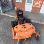 Orange SCAG commercial mower parked on a driveway outside a brick building, near a white door and an Amazon sign on the wall.