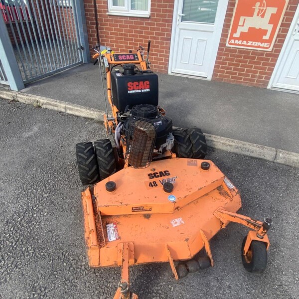 Orange SCAG commercial mower parked on a driveway outside a brick building, near a white door and an Amazon sign on the wall.