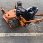 Orange riding lawn mower with exposed engine and large rear tires parked on asphalt surface.