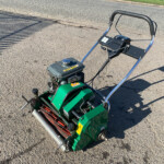Gas-powered garden tiller with a black handlebar resting on a paved road; engine and controls visible on the machine.