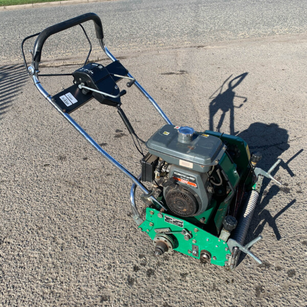 Green and black agricultural cultivator with a small gas engine mounted on a paved road, handlebars and controls visible.