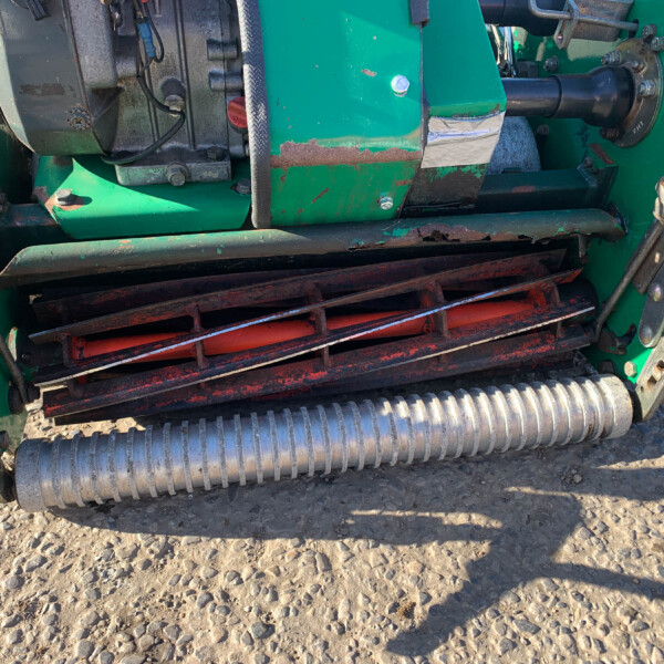 Close-up of a green farming machine with metal rollers and drive components on a dry, rocky field; visible rust and wear on the metal surfaces.