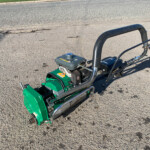 Green compact garden tiller/cultivator with a gray engine and curved handlebar on a paved surface, photographed from above.
