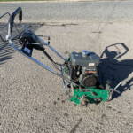 Gas-powered garden tiller with green frame and chrome handlebars resting on a paved surface outside.