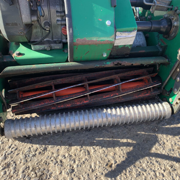 Close-up of a green agricultural machine with rusted metal plates, black rollers, and exposed mechanisms on a dirt surface.