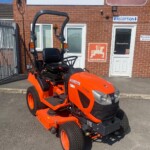 Orange Kubota ride-on lawn mower parked outside a brick building with reception signs and a large Russell Signs banner behind it.